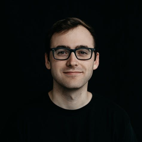 Headshot of a white male adult wearing glasses and a black t-shirt, in front of a black backdrop.